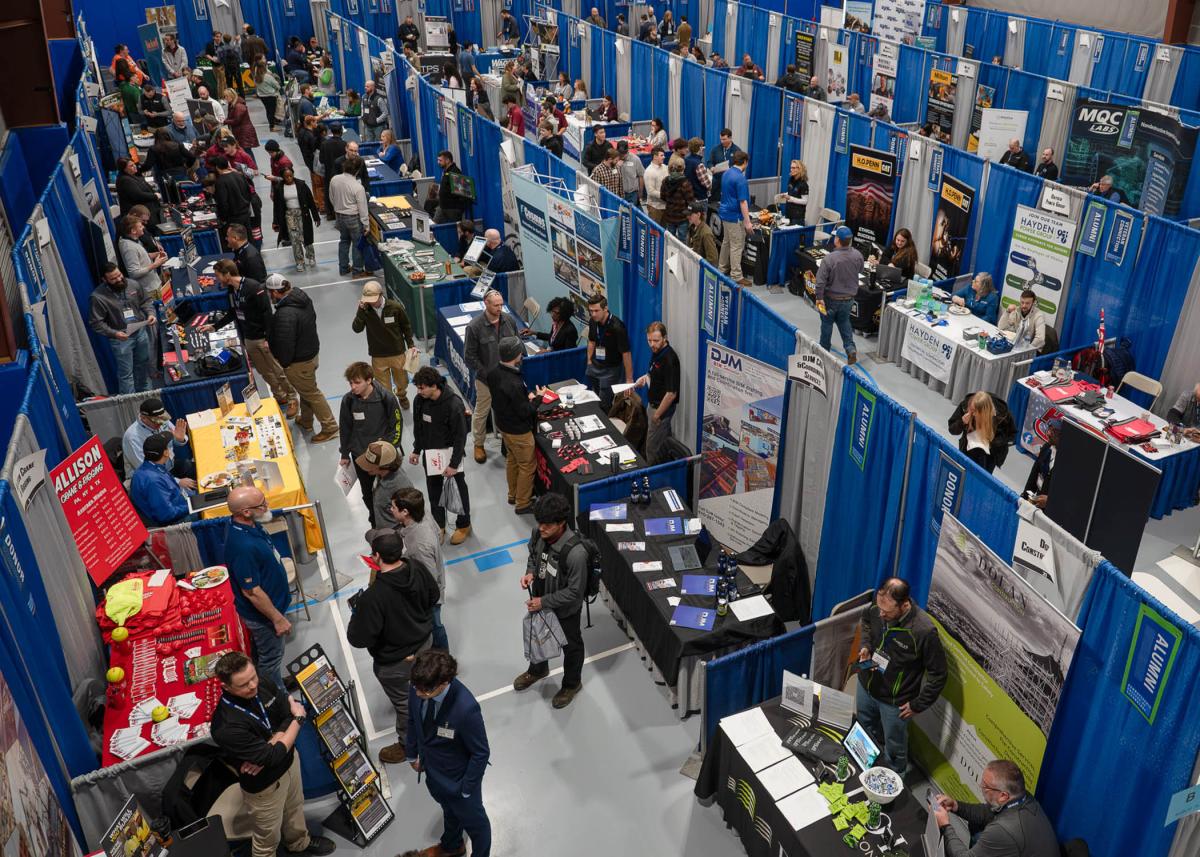 Overhead view of a gym filled with several rows of employer booths. Students walk among the booths and talk with the employers.
