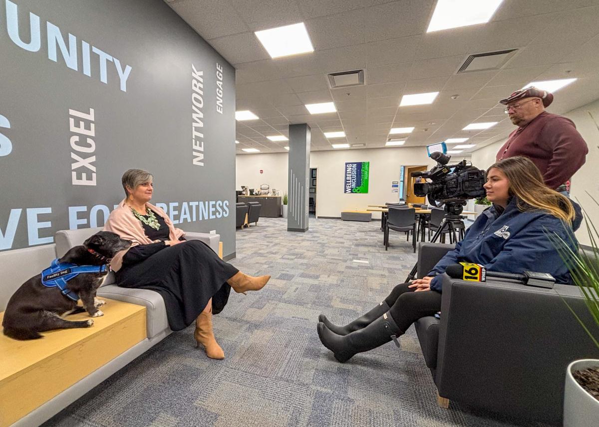 In Penn College's Hudock Center for Academic Excellence, a small dog sits on an end table, a wom sits on a comfortable chair next to the dog. A reporter sits in a chair opposite them, and a man with a camera stands next to the reporter.