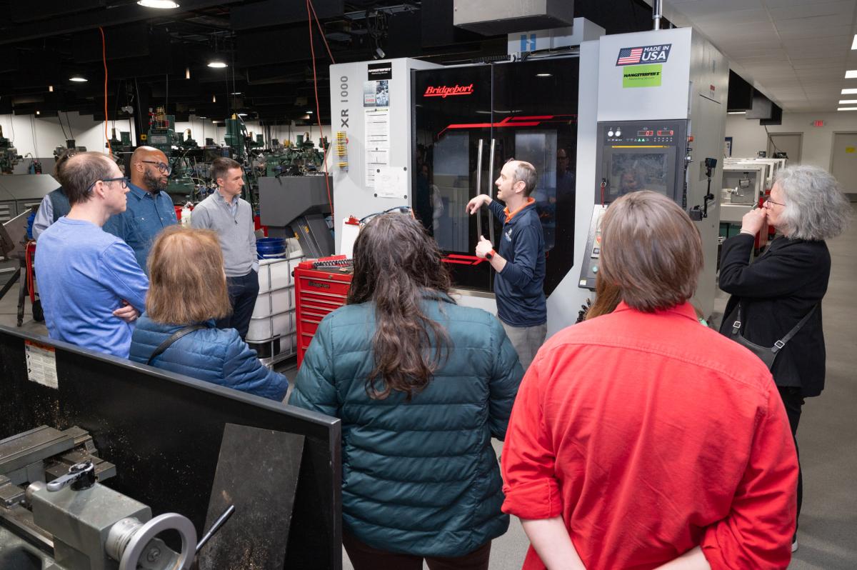 A man standing in front of a vertical machining center speaks to a group.