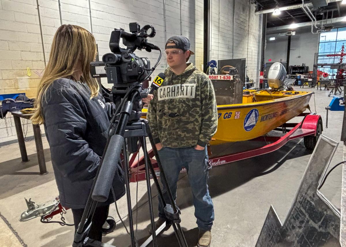 Inside Penn College's welding lab, a woman with a camera and microphone interviews a student who is standing near a bright yellow boat on a trailer.