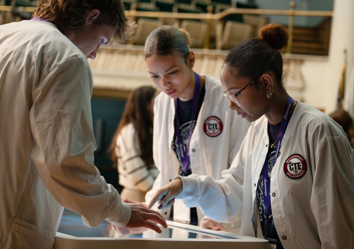 On the stage in Penn College's Klump Academic Center Auditorium, high school students in white lab coats stand around a large touchscreen table, tapping on a 3D anatomical image.