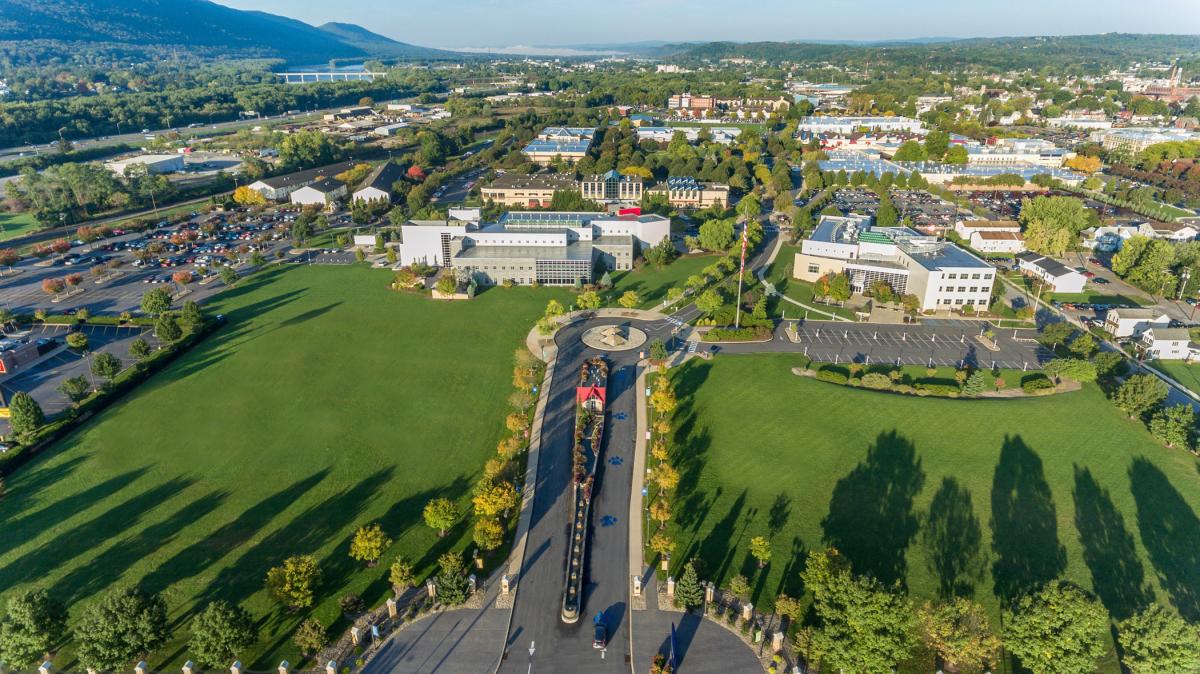 Aerial view of the campus entrance with lawns and a tree-lined street in the foreground and buildings in the background.