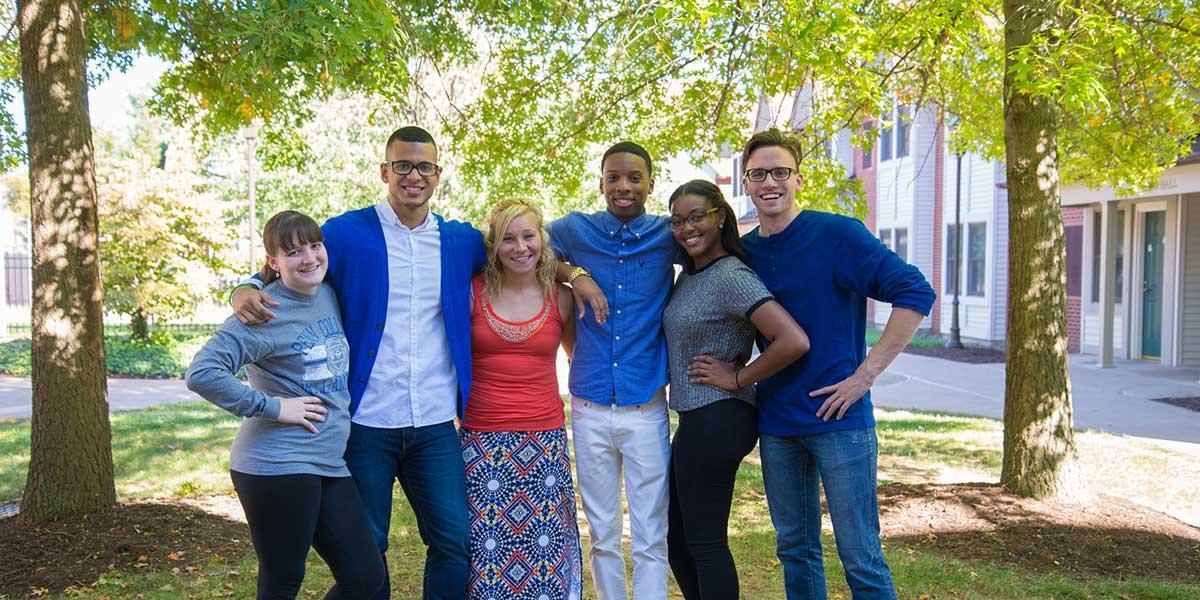 Six students pose with arms across shoulders under trees outside the Village.