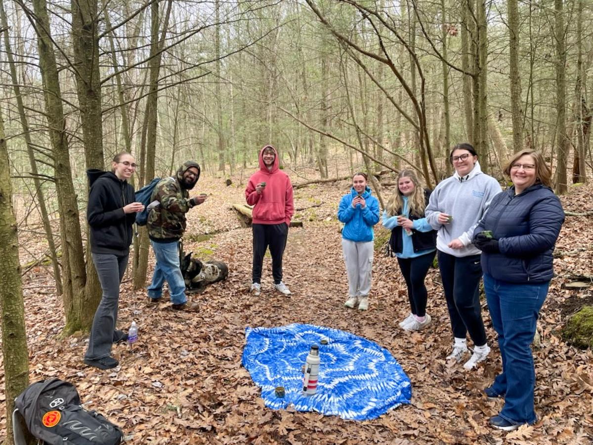 Students stand on a wooded trail holding small mugs. On the ground is a thermos.
