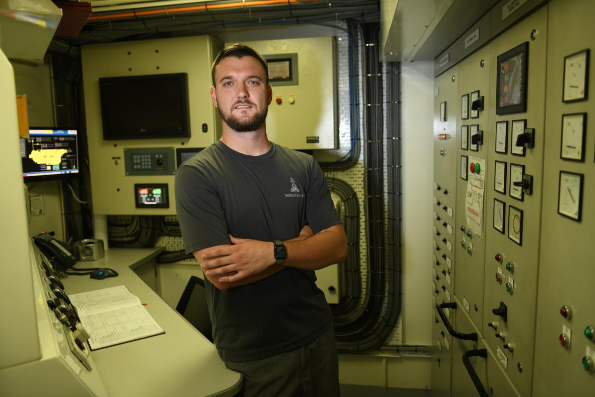 Person standing in an industrial control room surrounded by electrical panels and equipment.
