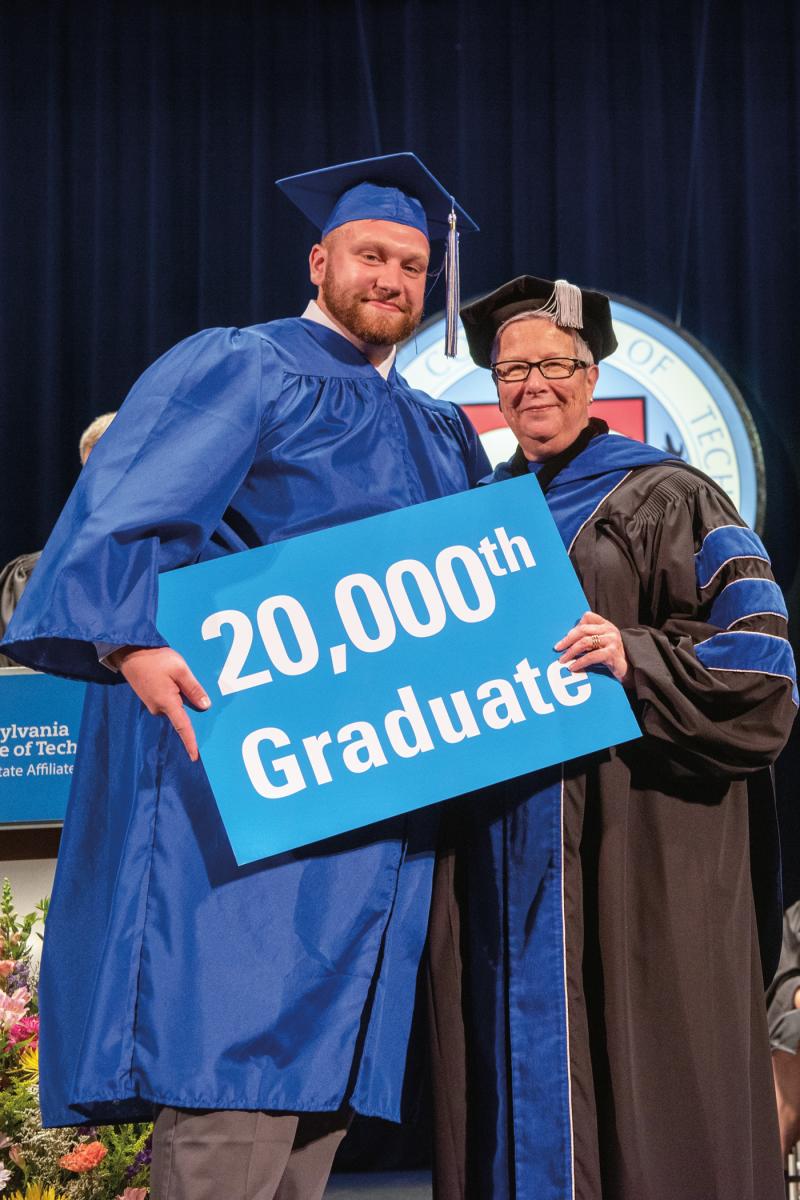Graduate holding a sign reading “20,000th Graduate” while standing with a faculty member on stage.