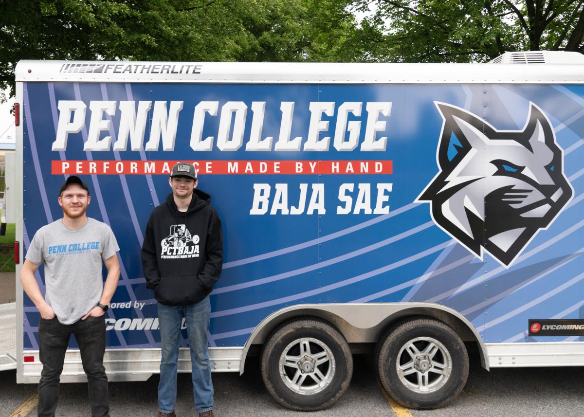 Two people standing in front of a Penn College Baja SAE team trailer.