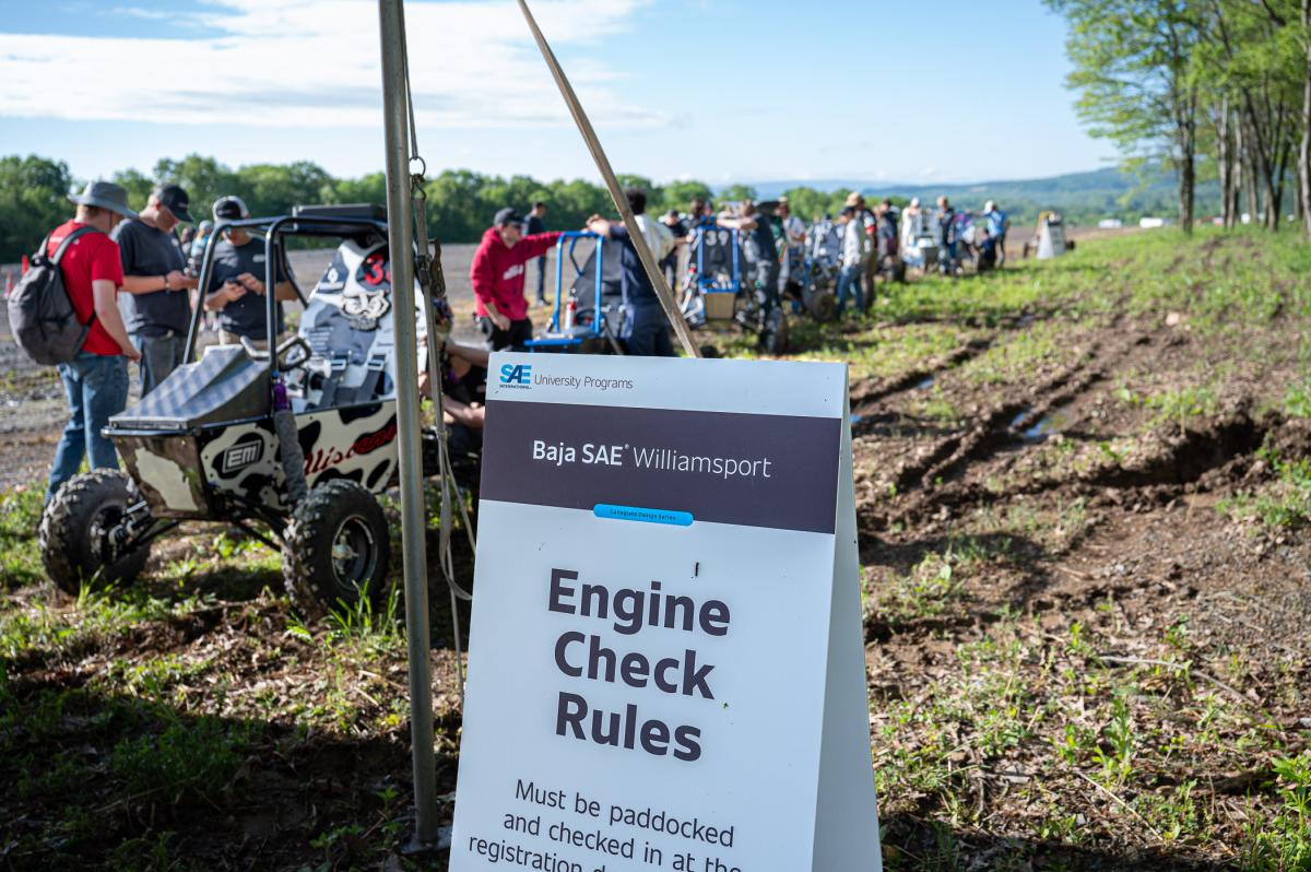 Sign reading “Baja SAE Williamsport Engine Check Rules” beside off-road vehicles lined up outdoors