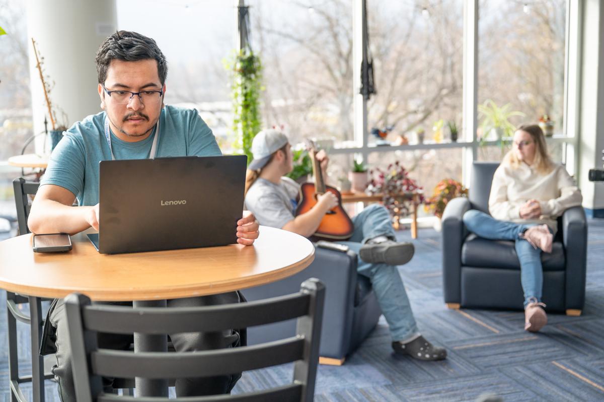 A student on a laptop sits at a high table while two students—one playing a guitar, the other watching—sit on lounge chairs behind in a glass walled commons area