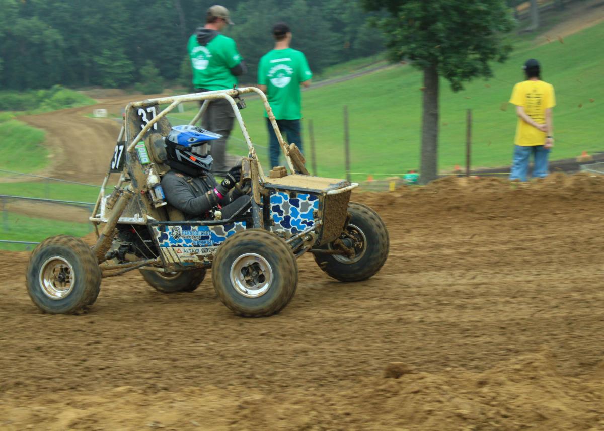 Off-road Baja vehicle racing on a muddy dirt track with spectators nearby
