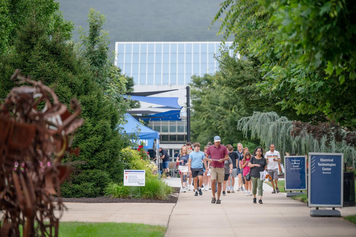 A crowd walk down the Mall among the trees, directional signs, and information tent for a summer Open House.