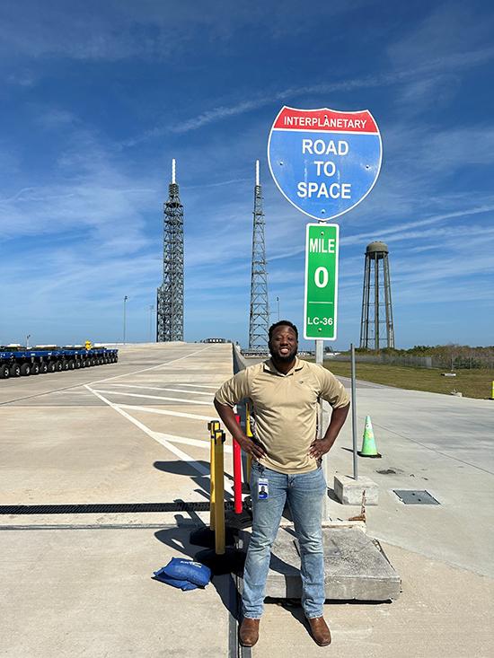 Dwight Alexandar stands under a sign that says 'Road to Space.' Behind him is a rocket launchpad.