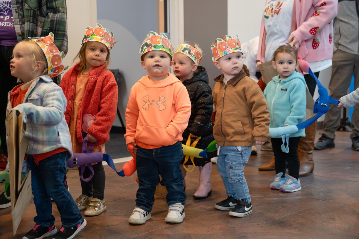 Toddlers wearing paper crowns standing in a line during a classroom activity or parade.