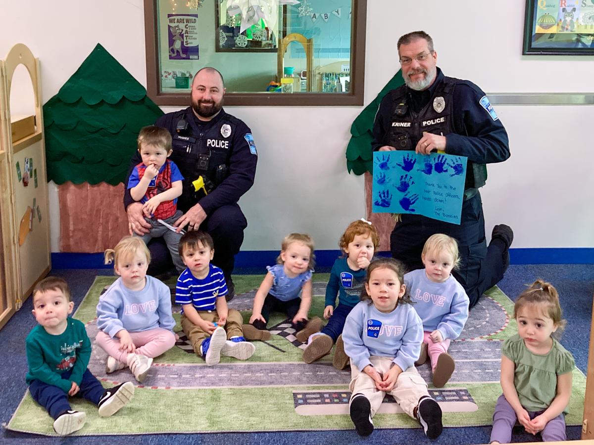 Police officers seated with toddlers in a classroom, holding a children’s artwork.