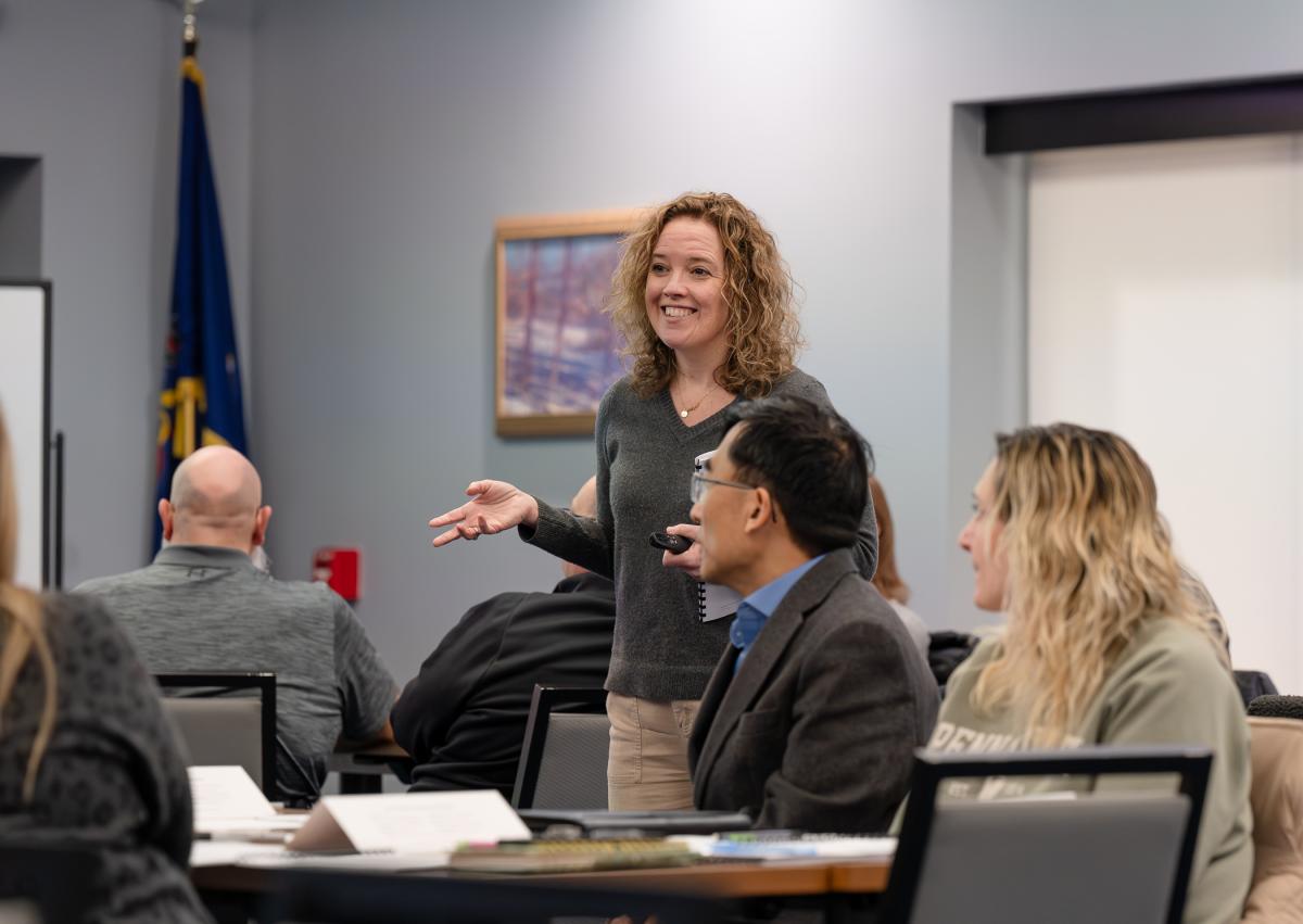 A trainer stands among tables of participants addressing the group.