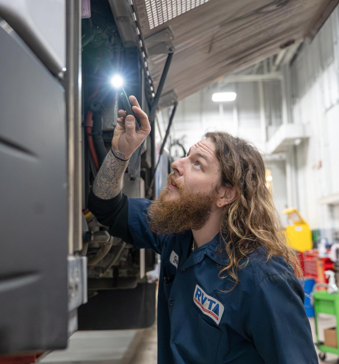 A Bus Mechanic Apprenticeship holds a small flashlight to inspect the inner workings of a bus.