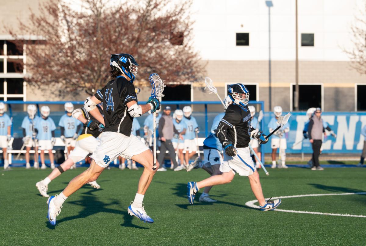 Lacrosse players running with sticks during a game on a turf field.
