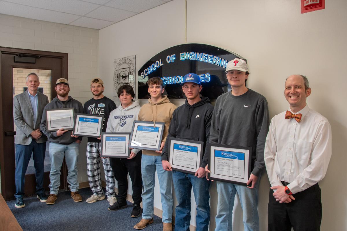 A group of Penn College students stand in an office holding framed certificates. The college president stands on one end of the group, and a dean stands on the other.