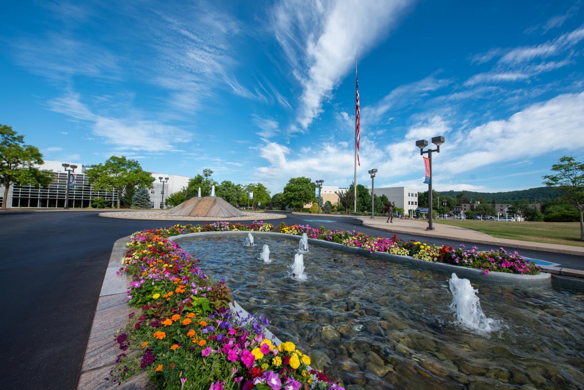 Campus fountain with flowers, flagpole, and buildings under a blue sky.