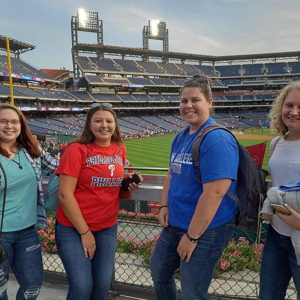 Four students lean against a fence with a baseball stadium in the background