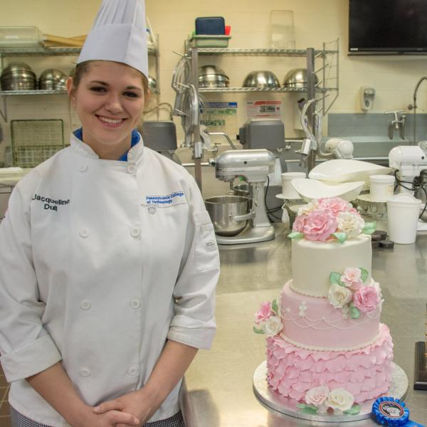 Chef standing beside a decorated three-tier cake in a baking lab.