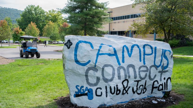 The Rock, a large boulder that serves as a billboard, is painted white with the words "Campus Connect: Club & Job Fair" in blue, gray and black. Small logos for various student organizations are painted around the words.