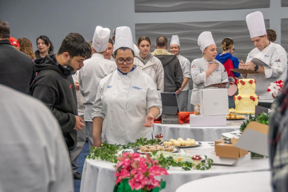 People gather around tables in Penn College's Thompson Professional Development Center. Near the center of the photo, a student in a chefs uniform gestures toward pastries on her table while talking with a young man.