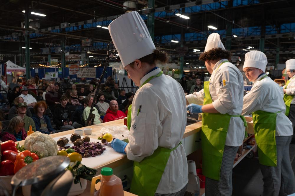 Students in chefs uniforms with green aprons prepare food on the Pennsylvania Farm Show’s Culinary Connection stage. Various vegetables are arranged on the counter. A seated audience watches the cooking demonstration in a large indoor venue with booths and banners in the background.