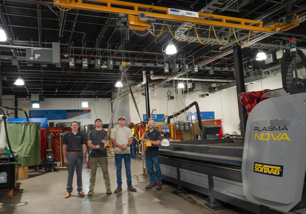 Four people standing in Penn College’s Lycoming Engines Metal Trades Center beneath an overhead crane, surrounded by welding equipment, fabrication tools, and a plasma cutting machine.