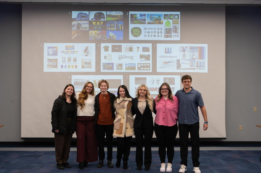 Penn College graphic design students stand in front of a large projection screen that shows logos and other branding materials.