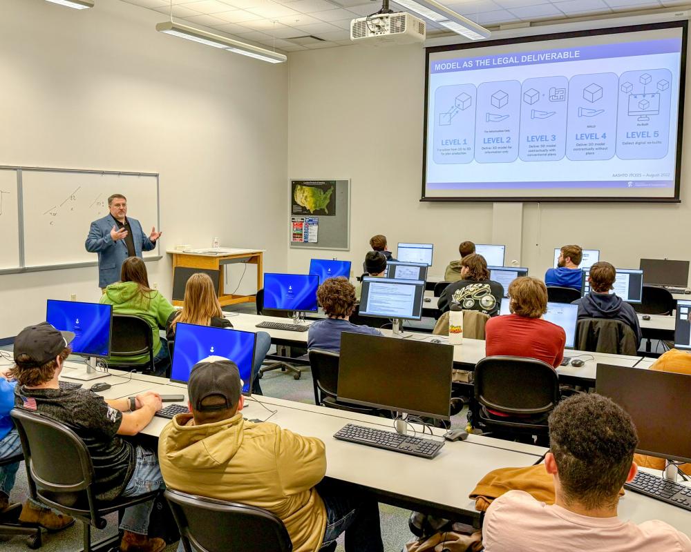 A person speaks in front of a classroom outfitted with a computer for each student and a large projection screen.