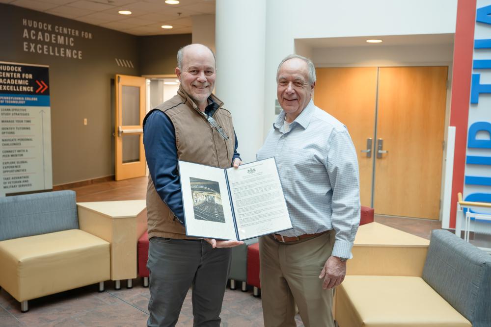 Two men stand side by side. One is holding an award.