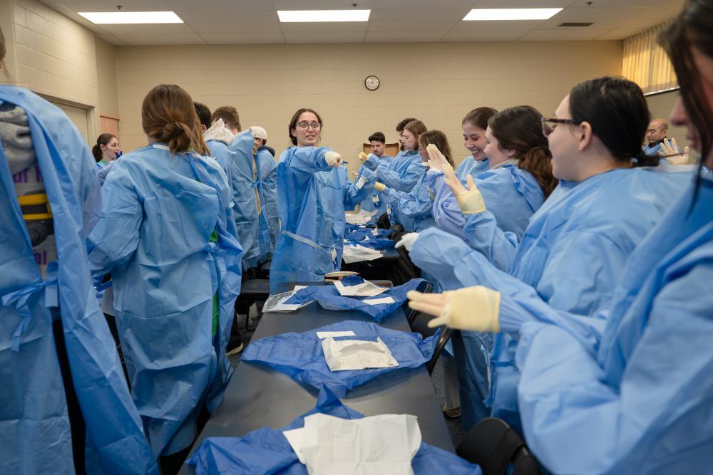 A recent alumna points while demonstrating a room full of students how to properly put on gowns and gloves.
