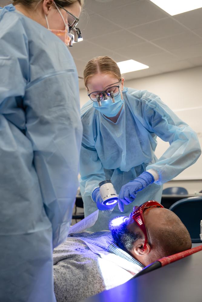 A dental hygiene student in protective medical gear shines a blue light into a patient’s mouth while another student observes.