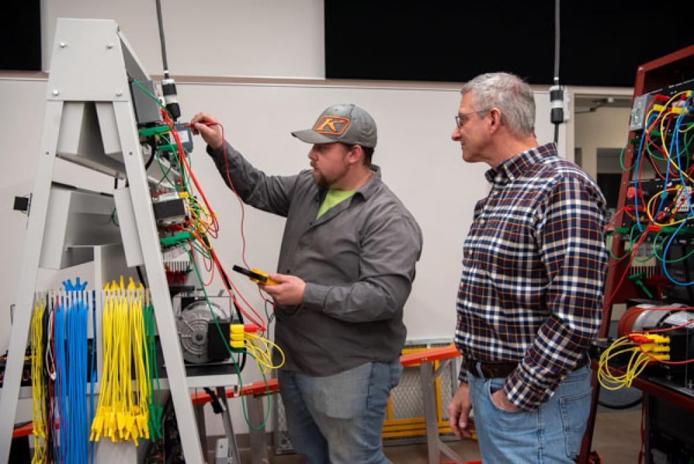 A faculty member observes an apprentice working on a mechatronics control panel.