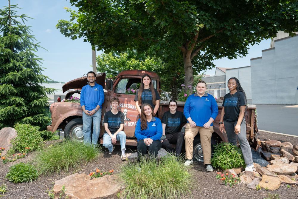 Group of students and staff posing in front of a vintage truck on a landscaped college campus.