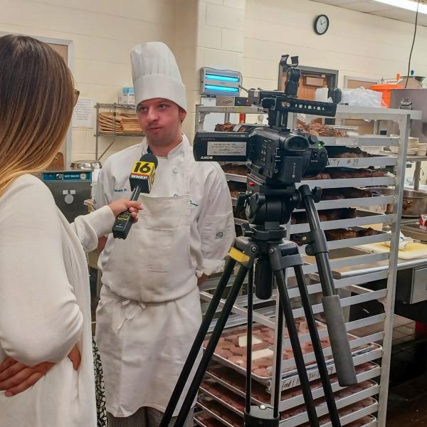 In a commercial kitchen, a woman holds a microphone up to a student wearing a chefs jacket and toque. A TV camera is on the right.