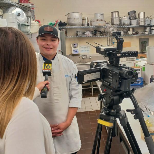 In a commercial kitchen, a woman holds a microphone up to a student wearing a chefs jacket. A TV camera is on the right.