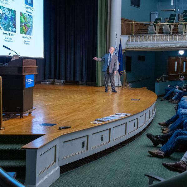 Mark Shiflett, wearing a suit and tie, stands on the stage of the Klump Academic Center Auditorium and gestures toward a larger projection screen while students watch from auditorium seats.
