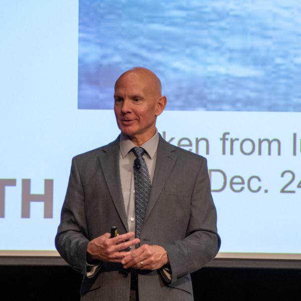 Mark Shiflett, wearing a suit and tie, stands in front of a large projector screen.