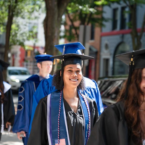 Students in graduation caps and gowns walk in a line down down a street toward the Journey Bank Community Arts Center.