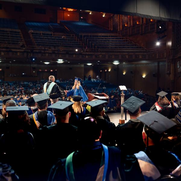 A graduate in a blue graduation cap and gown shakes the hand of Penn College President Michael J. Reed.