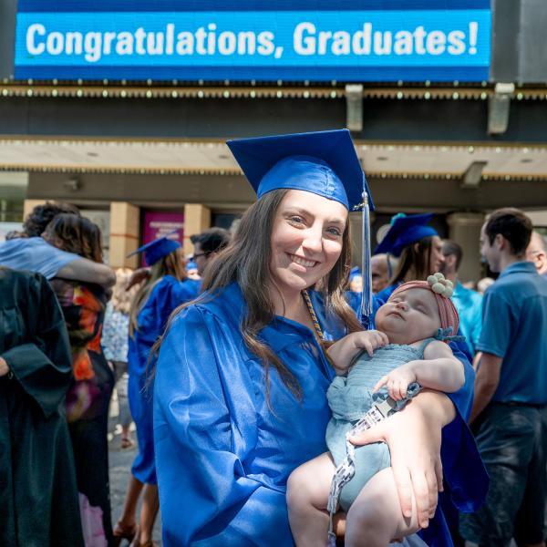 A woman in a blue graduation cap and gown holds a sleeping infant in front of the Journey Bank Community Arts Center marquis, which says "Congratulations, Graduates!"