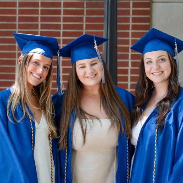 Three women in white dresses and blue graduation caps and gowns stand together.