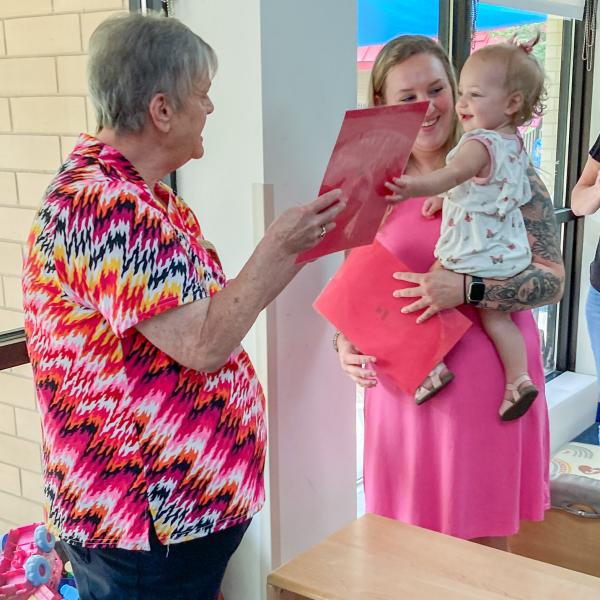A grandmother hands a book to a smile child. The child is being held by another adult.