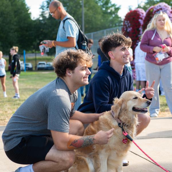 Students kneel on either side of a dog and look at a camera.