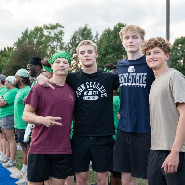 Four students with arms around one another look at the camera, one giving "victory" sign with his fingers. In the background, their teammates line the sideline of UPMC Field.