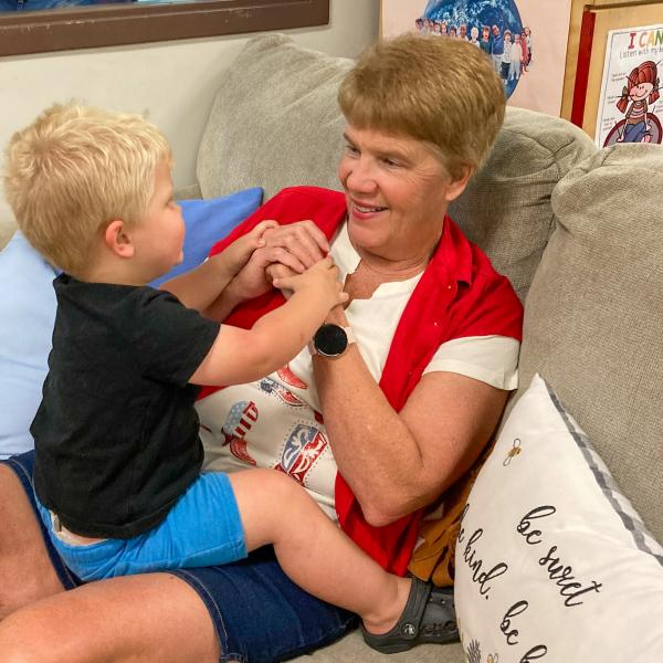 A toddler looks at his grandmother while holding her hands and sitting on her lap on a couch.