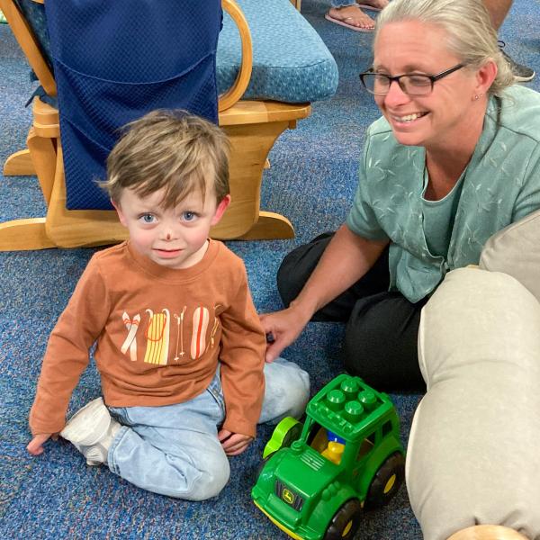 A grandmother smiles while her grandson plays with a toy John Deere tractor.
