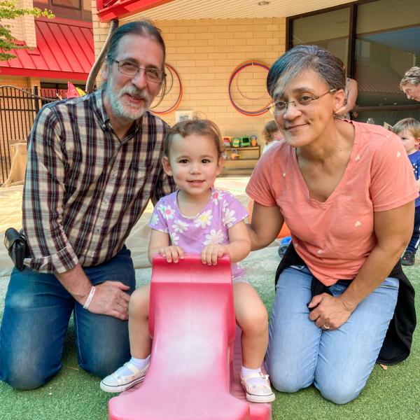 A grandfather and grandmother kneal next to a child on a rocking toy.